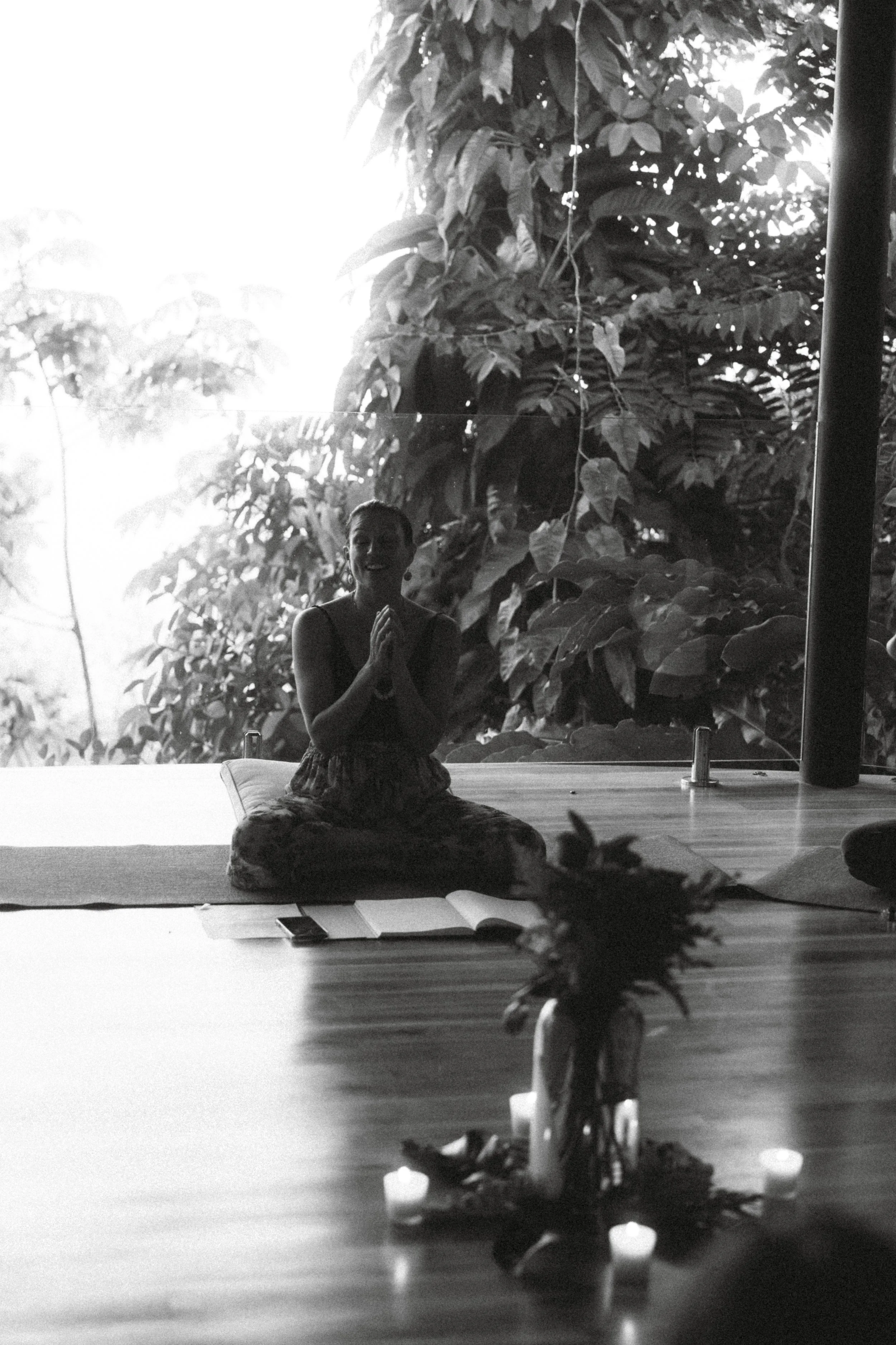A woman practicing yoga in a seated prayer pose in front of a large window with lush greenery outside. There are candles and a small plant on the table nearby.