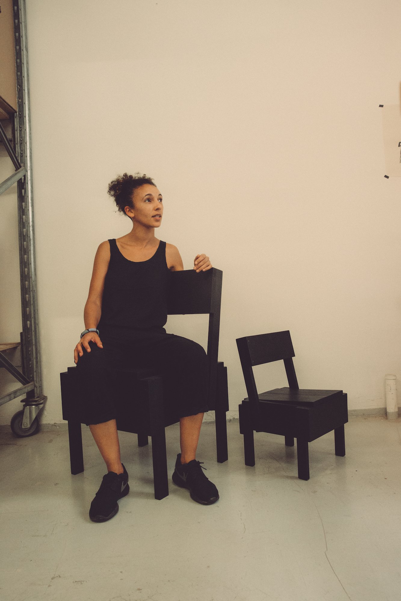  Corinna Sy, a designer and co-founder of Cucula, sitting on the black edition of Sedia Uno from Autoprogettazione – a piece designed by Enzo Mari and made by Cucula. Next to the Sedia Uno is the Bambinooo chair, also designed by Mari. 