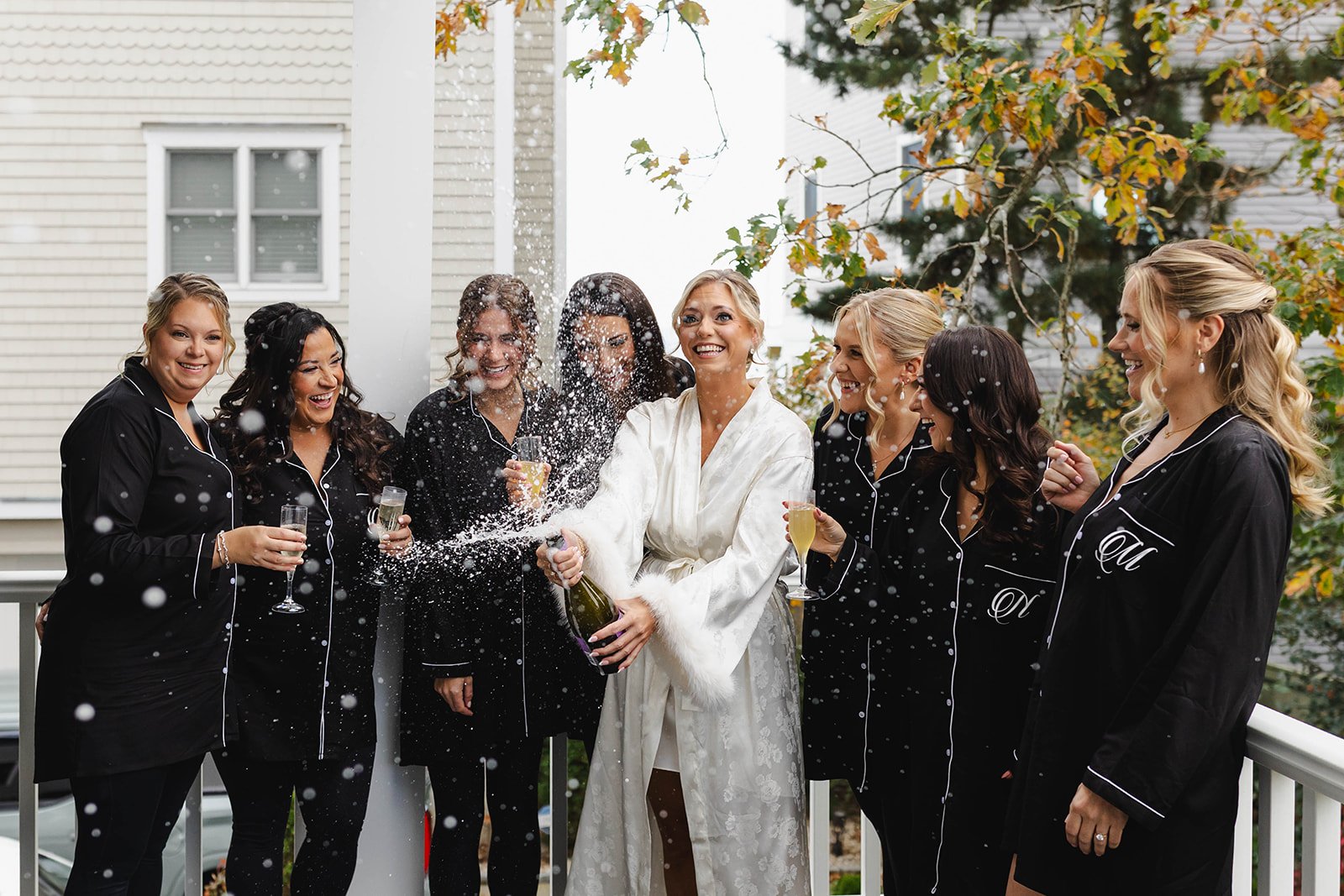 A bride in a white wedding gown celebrating with her bridesmaids and friends on a balcony, popping champagne and spraying beverage, all wearing matching black pajamas with white piping.
