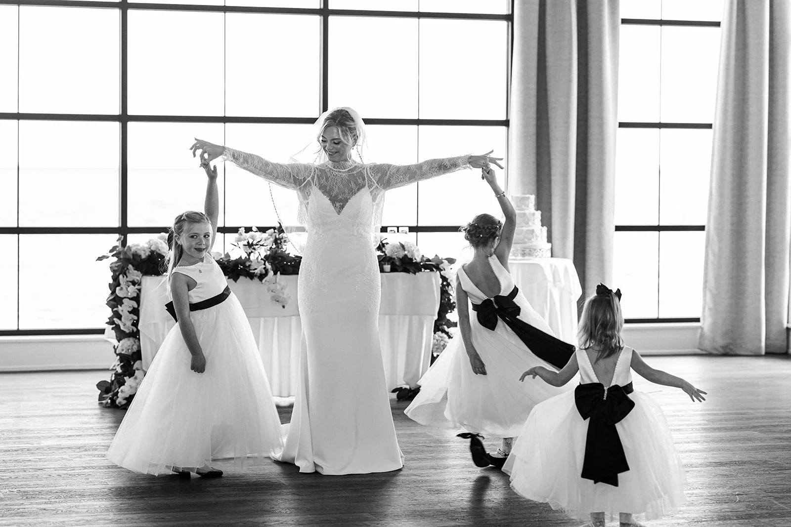 A bride and four young girls dancing at a wedding reception in front of large windows with curtains. The bride is wearing a wedding dress with lace sleeves, and the girls are wearing white dresses with black ribbons.