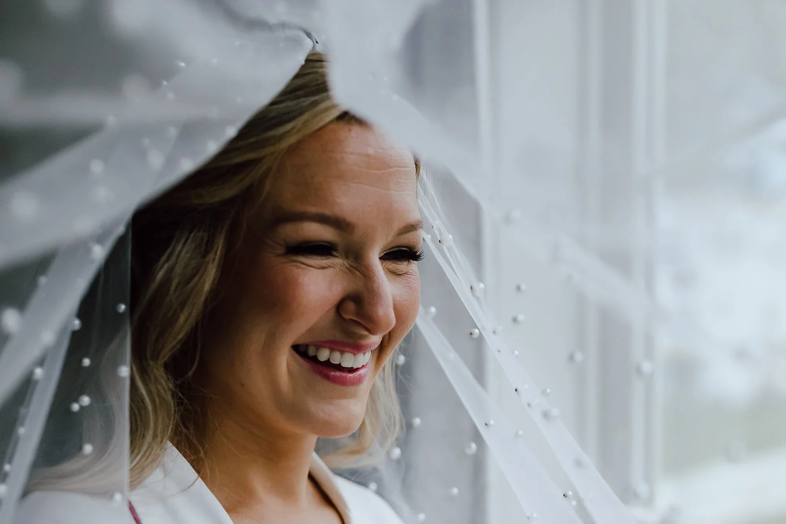 Smiling bride with a beaded veil near a window.
