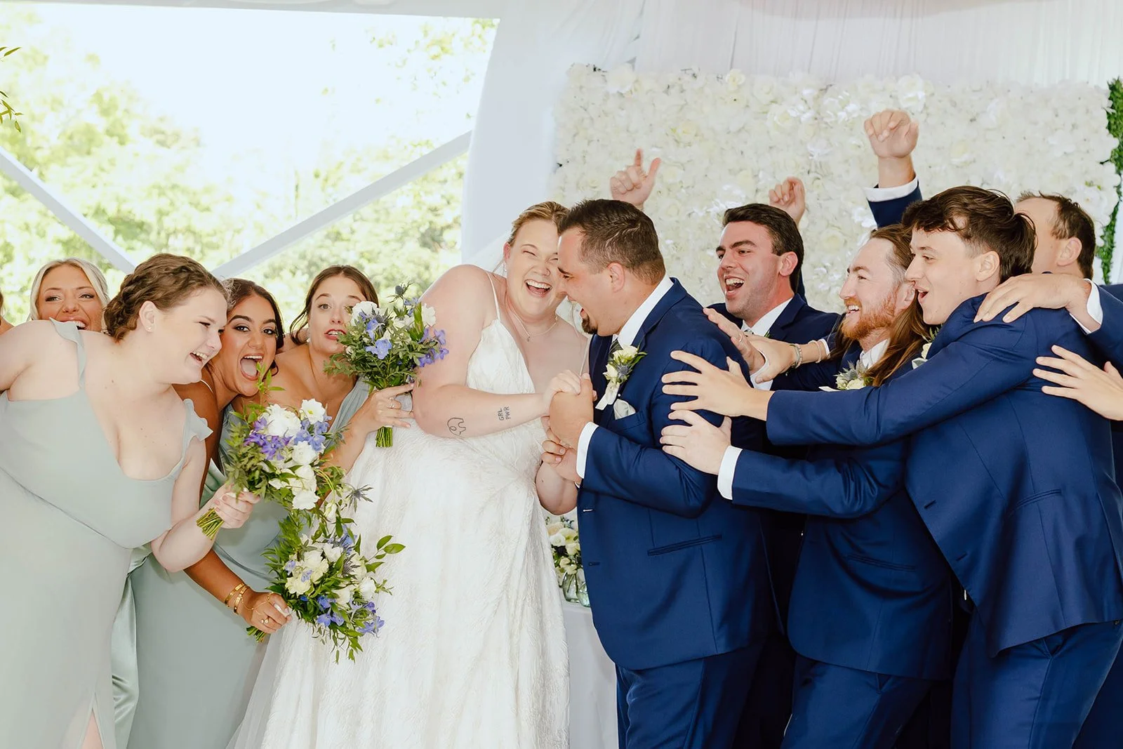 A group of bridesmaids and groomsmen celebrating at a wedding, with the bride and groom at the center, smiling and laughing.