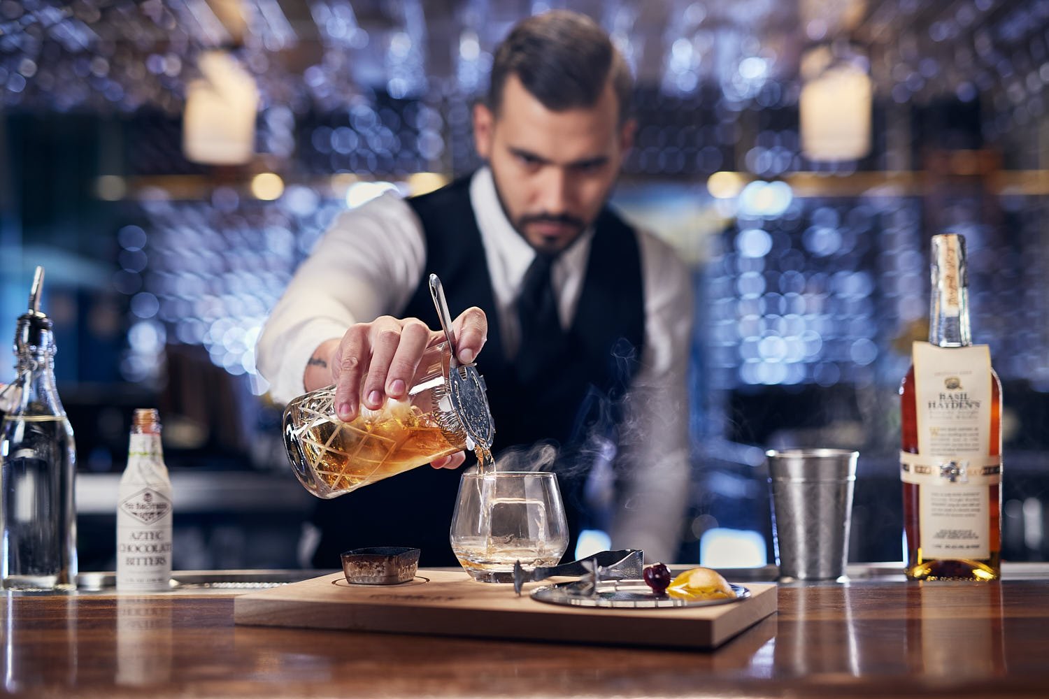 Lifestyle photo of a bartender with a white shirt, tie and black vest pouring a smoked old fashioned into a rocks glass with a large single ice cube at a restaurant bar. There is a bottle of Basil Haden Bourbon and small bottle of bitters on the bar 