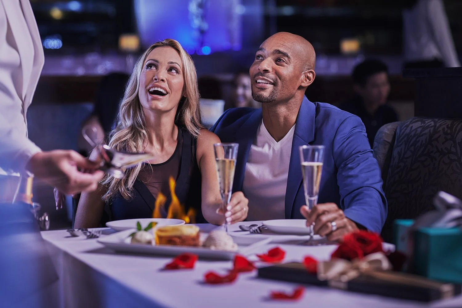 A glamorous couple at a table at Eddie V's restaurant with a server pouring flaming rum sauce onto bananas foster dessert. Romantic restaurant setting with rose petals and champagne on the table. Seattle restaurant photography.
