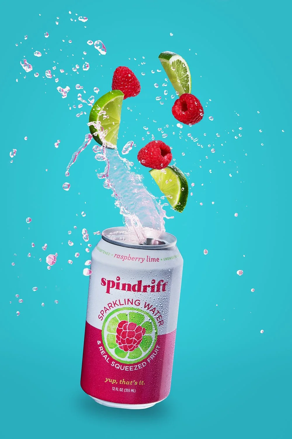 A can of Spindrift water splashing water out of the top.  Limes and raspberries are above suspended above the can. On a blue background. Bright crisp lighting. Seattle CPG packaging photographer.
