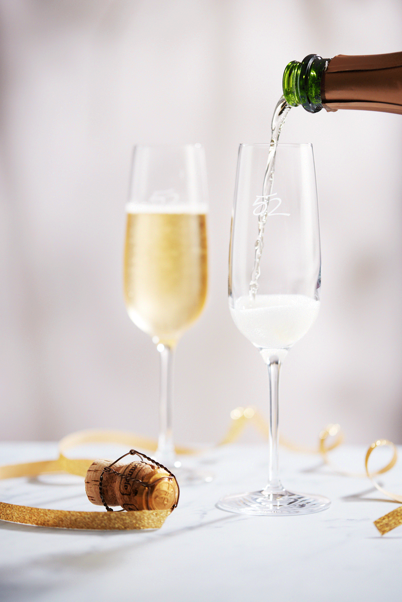 Bottle of sparkling champagne pouring into a champagne flute on a white marble table surface. Also on the table are a gold holiday ribbon and the cork. Created by Food & Beverage Photographer and Director Mike Gluckman.