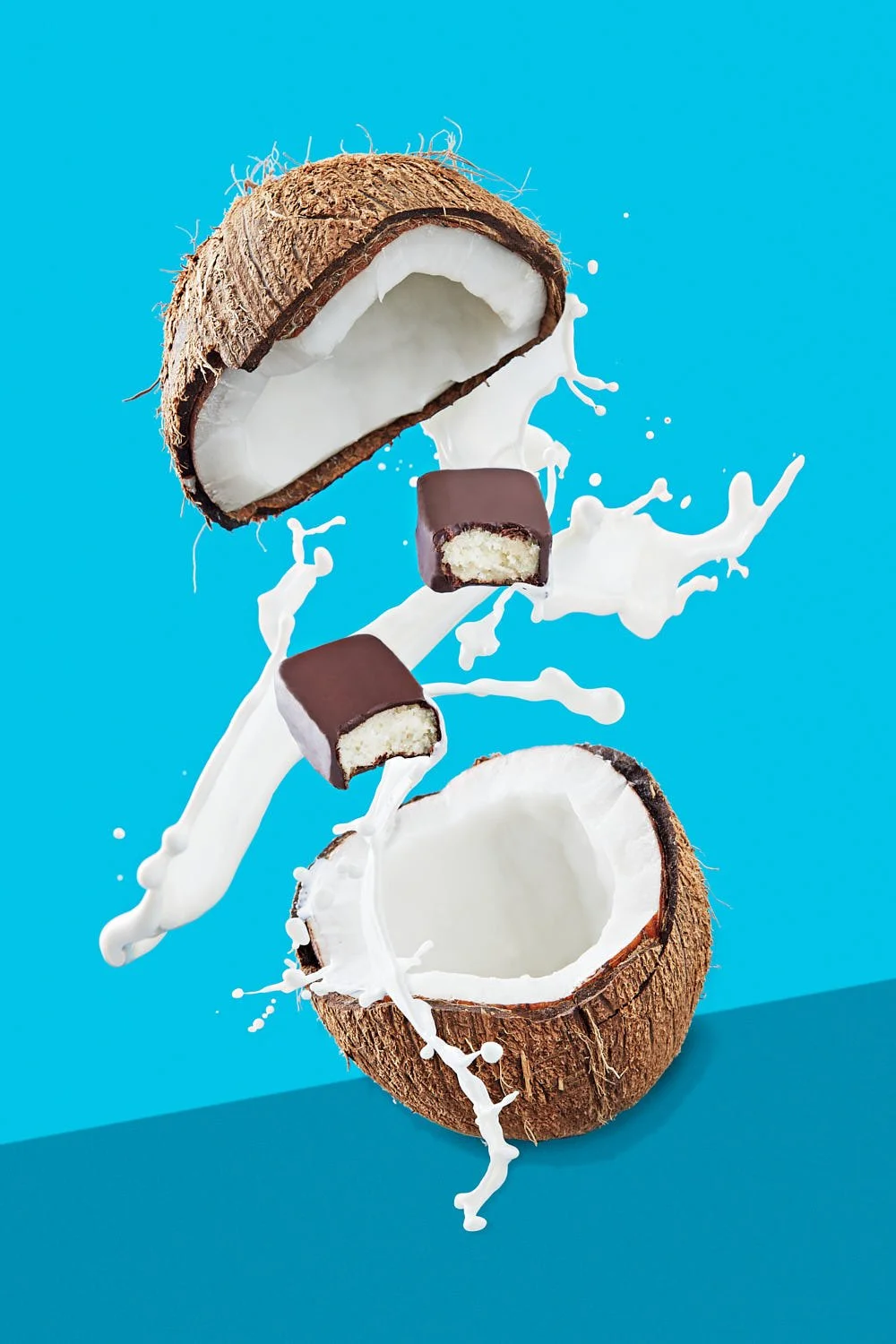 Two  chocolate bars and a splash of coconut milk in between the halves of a split coconut on a blue background. Seattle CPG packaging photography.