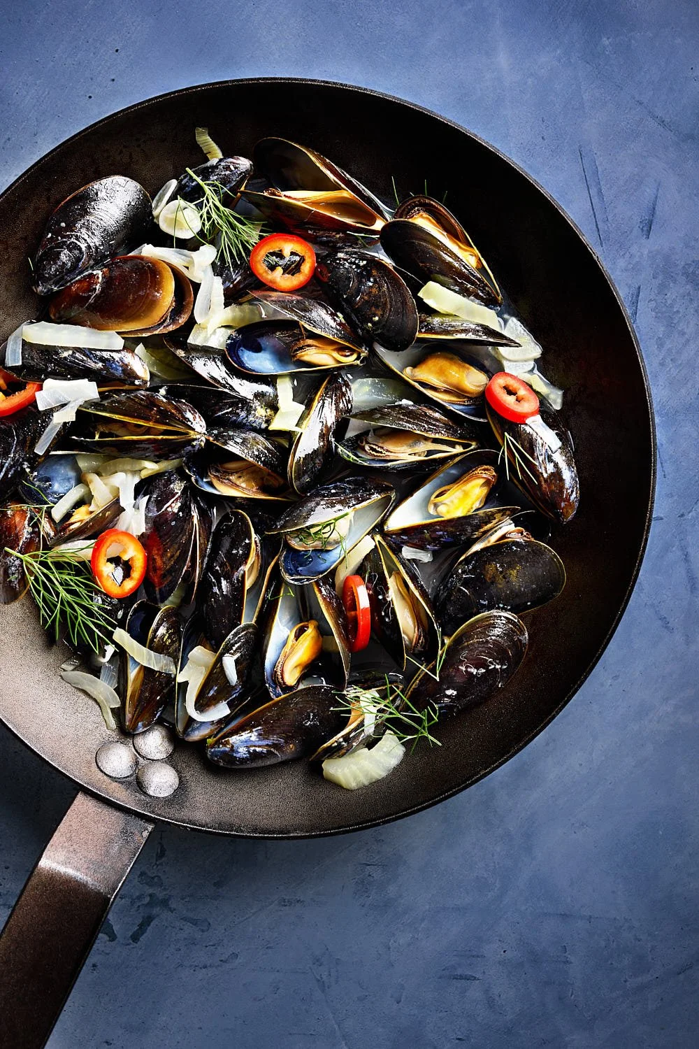 Mussels, fennel, onions and red chilis in a carbon steel sauté pan on a blue surface. Soft bright lighting. Editorial feel. Created by Seattle based food photographer Mike Gluckman.
