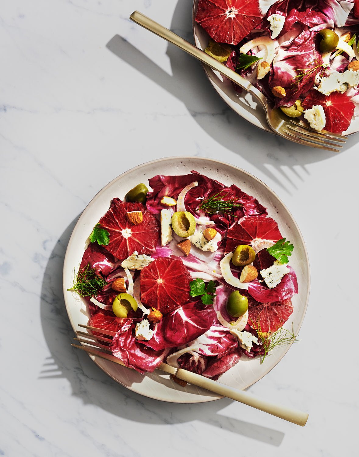 Overhead of two plates of radicchio salad with blood orange and green olives on a marble surface with gold forks. Crisp shadows and bright lighting. 