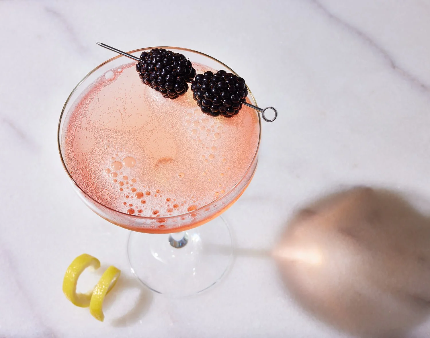 Blackberry champagne cocktail in a coupe glass and garnished with a lemon twist and two blackberries on a skewer. The drink is on a marble table. Bright poppy lighting with caustics and a strong shadow from the glass. Mike Gluckman is a Seattle drink