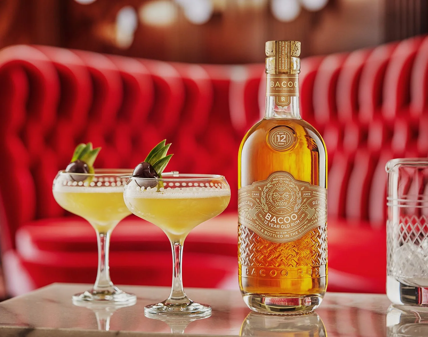 Beauty shot of a bottle of Bacoo rum sitting on a marble table with two Mary Pickford cocktails in fancy coupe glasses. Location is a swanky cocktail lounge with a red velvet couch. Cocktail photography in Seattle. Seattle drink photo studio.