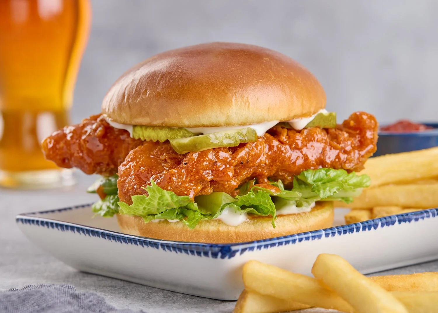 Close-up of a hot honey fried chicken sandwich with pickles, lettuce, and mayonnaise on a bun, served with French fries and a cold beverage in the background.