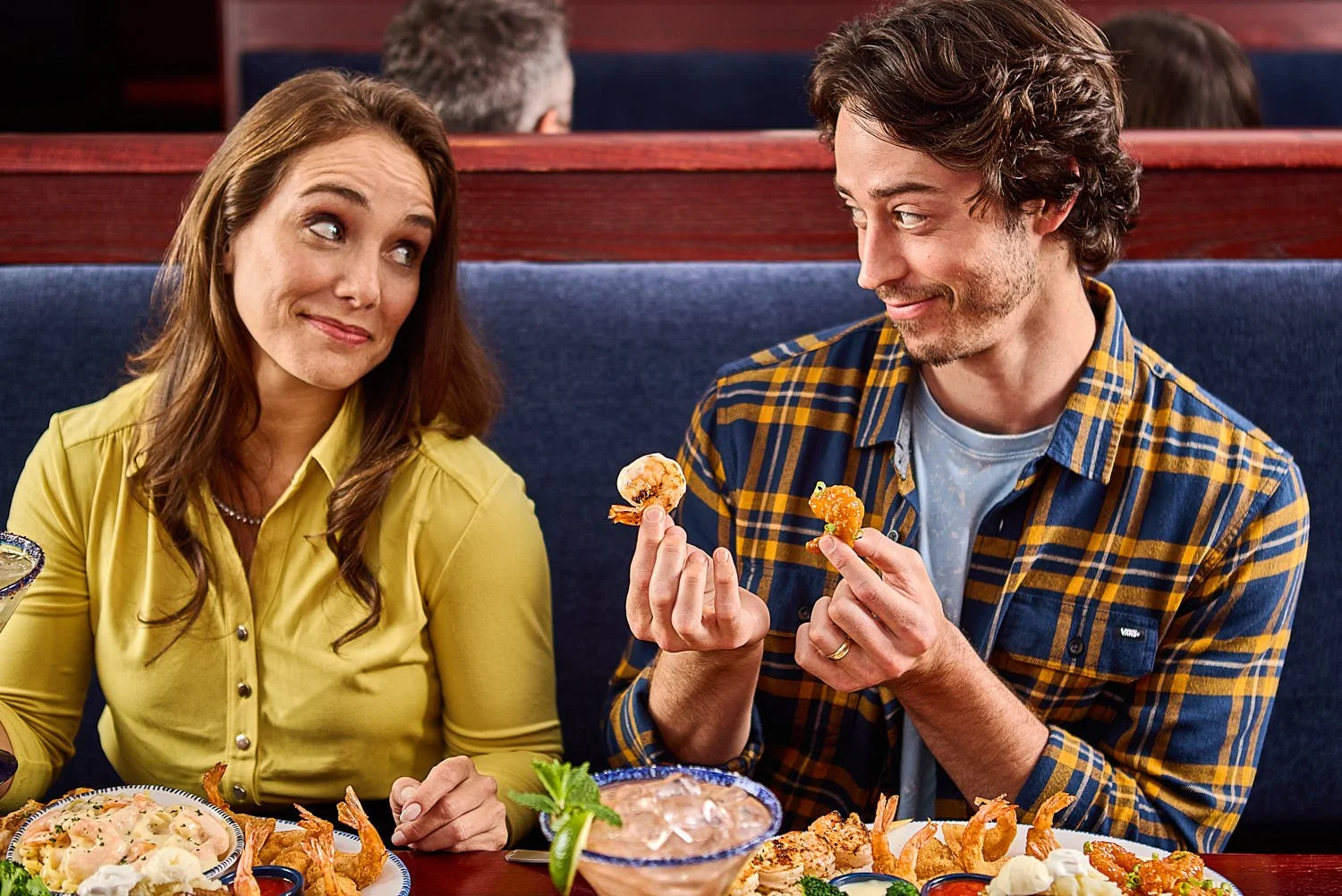A 25-35 year old man and woman sitting in a restaurant booth. He has a shrimp in each hand and they are giving each other a funny look. Photographed by Seattle restaurant lifestyle photographer Mike Gluckman.