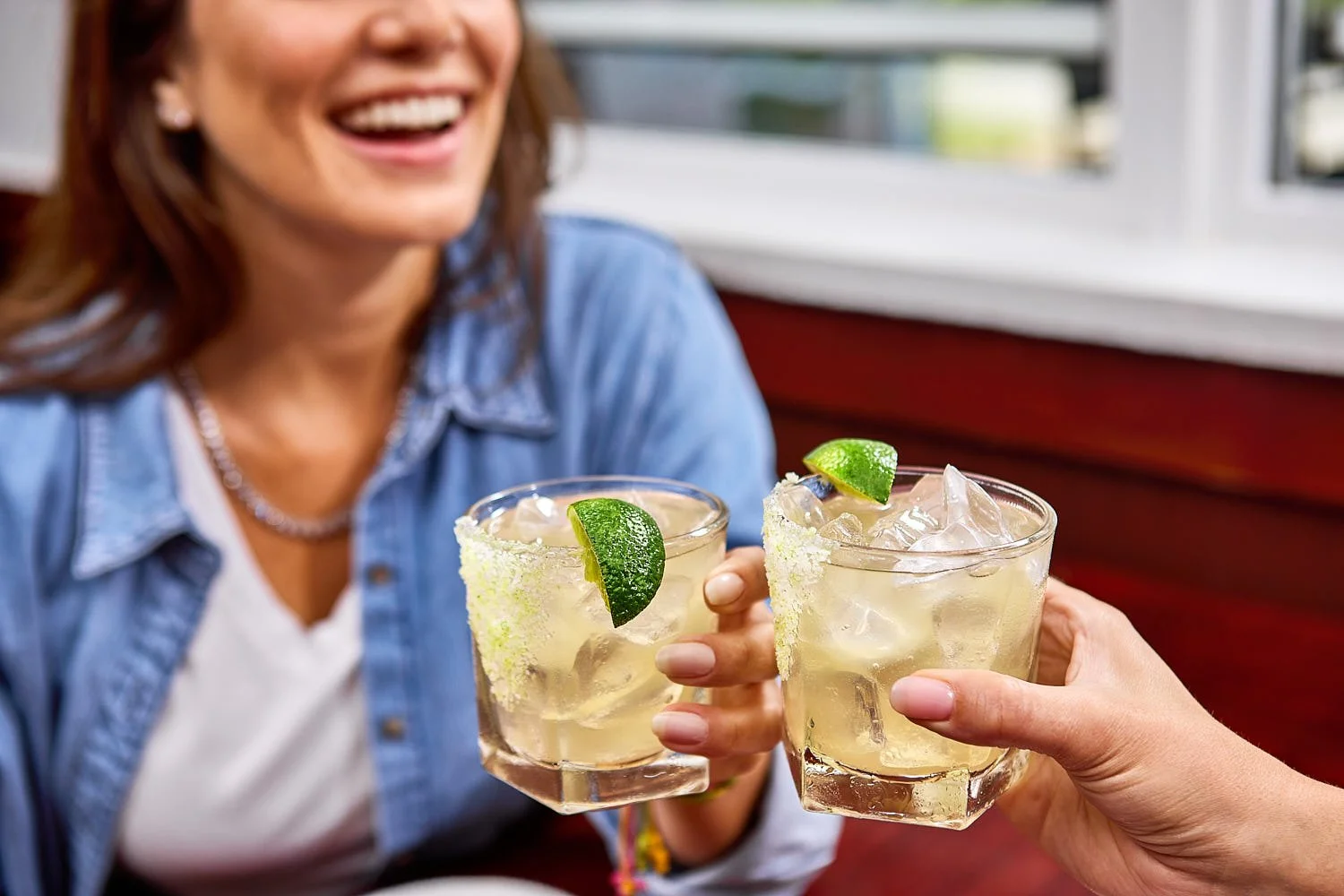 Two women toasting Margarita’s in a restaurant. Shot is close on cocktails showing one woman in a blue shirt.