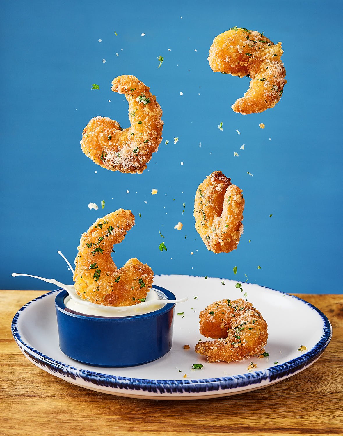 Fried shrimp being dropped into a bowl of creamy white dipping sauce with green herbs, with some shrimp and herbs mid-air above a white plate on a wooden surface against a blue background.