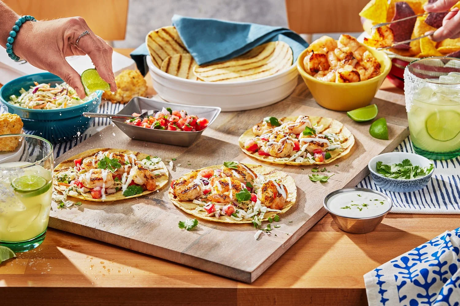 A dining room setting with shrimp tacos on a table along with shrimp, salsa, slaw and tortillas. There are margaritas in glasses and chips. A hand is squeezing a lime on to tacos. Bright high key lighting with sharp shadows. Photographed by Seattle b