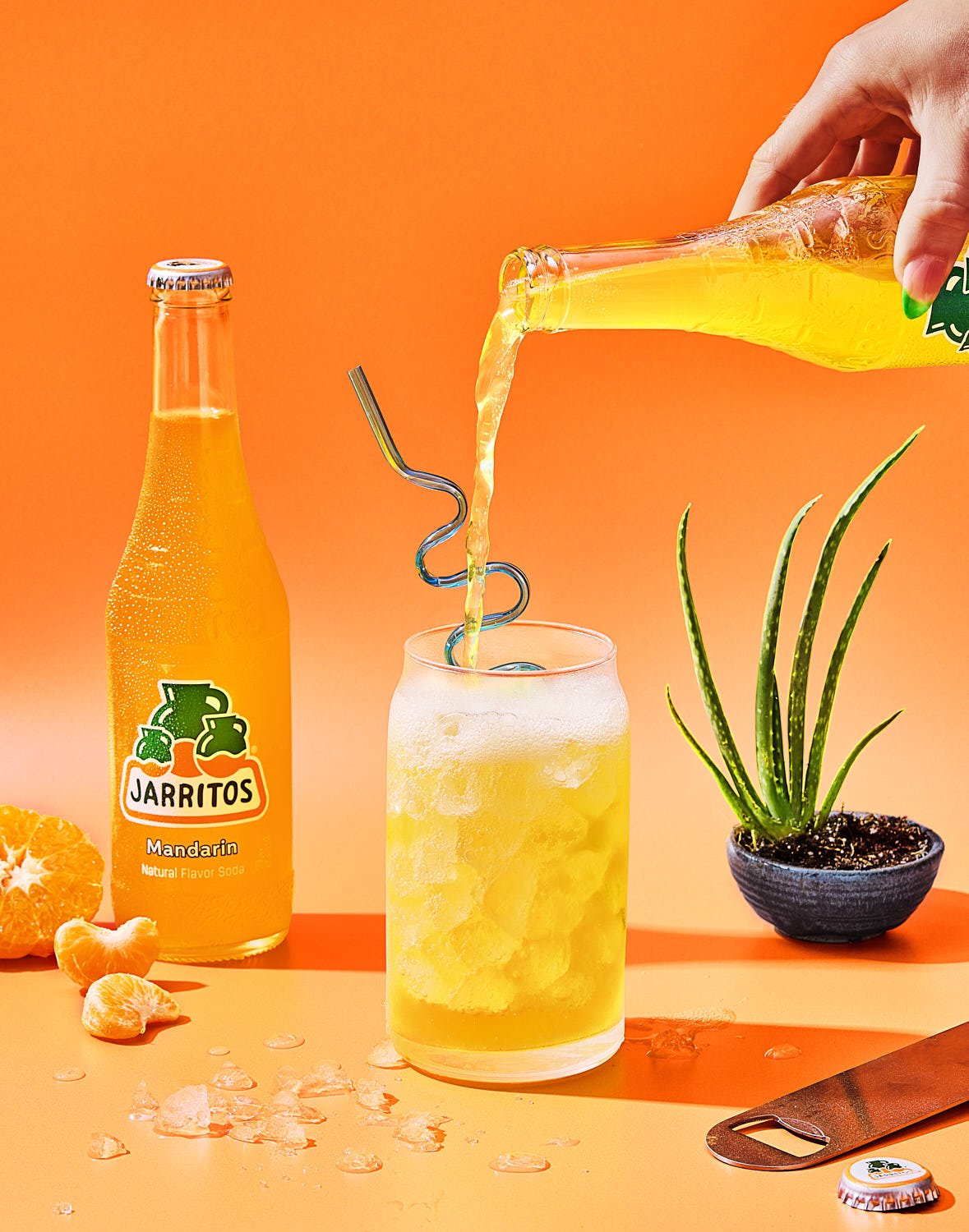 A hand pouring orange Mandarina soda from a glass bottle into a glass of ice, with another bottle and a sliced tangerine nearby, a small plant in a black pot, and a bottle cap on an orange surface against an orange background.