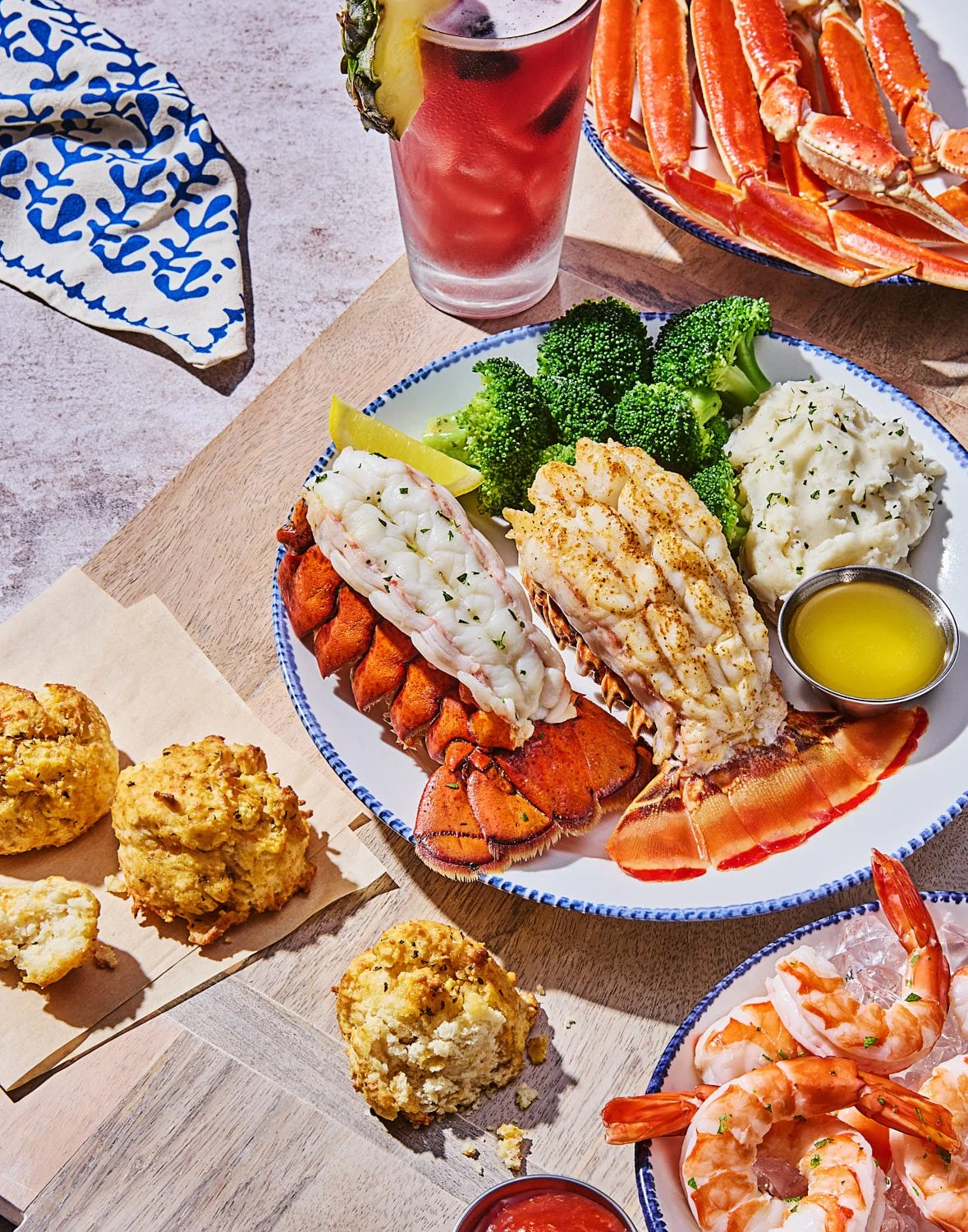 High angle beauty shot of a plate with two lobster tails accompanied by broccoli and mashed potatoes. Background is a large wood board with fresh baked Cheddar Bay biscuits on the side. There is also a plate with crab legs and a bowl of shrimp cockta