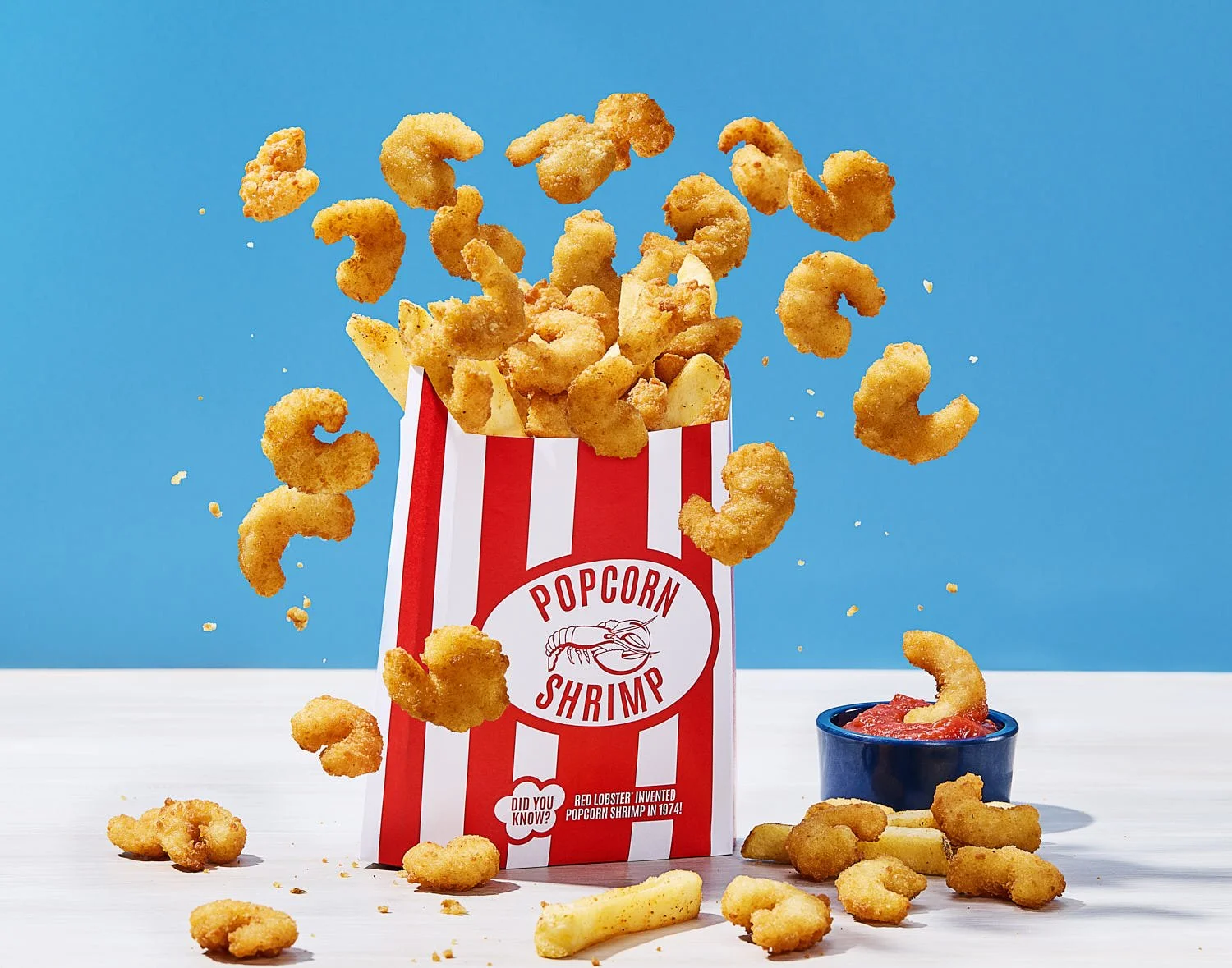  A red and white striped popcorn-style bag overflowing with golden, crispy popcorn shrimp and a few French fries. Some shrimp appear to be mid-air, as if popped from the bag. Next to it is a small blue cup filled with cocktail sauce, with one shrimp 