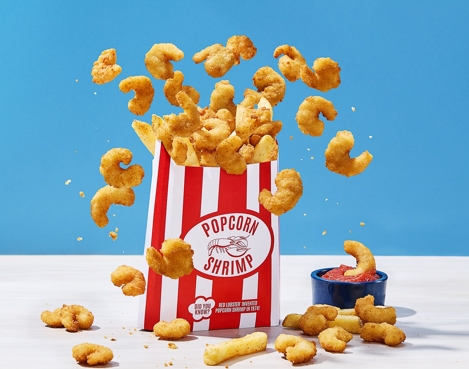  A red and white striped popcorn-style bag overflowing with golden, crispy popcorn shrimp and a few French fries. Some shrimp appear to be mid-air, as if popped from the bag. Next to it is a small blue cup filled with cocktail sauce, with one shrimp 