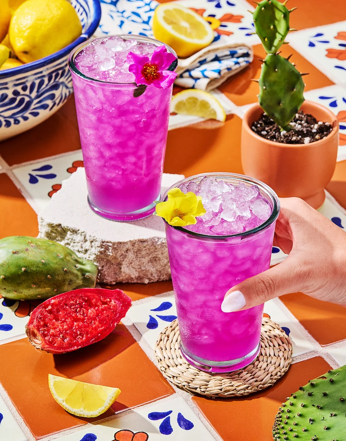  Two vibrant magenta aguas frescas filled with crushed ice, each garnished with a colorful edible flower, sit on a patterned tile surface. One glass rests on a woven coaster as a hand reaches for it, while the other stands on a stone block. Surroundi
