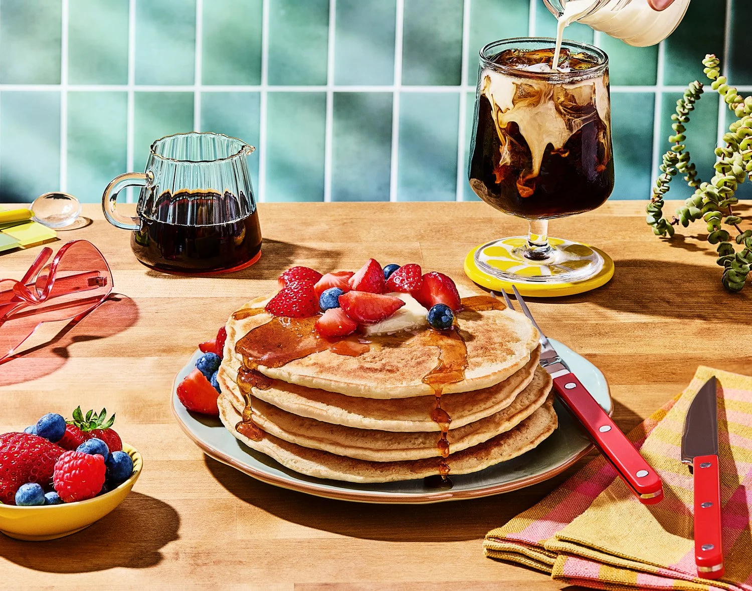 A sunny breakfast scene featuring a stack of pancakes topped with butter, syrup, strawberries, and blueberries on a plate with red-handled cutlery. A glass of iced coffee with cream being poured swirls beside the plate, accompanied by a syrup pitcher