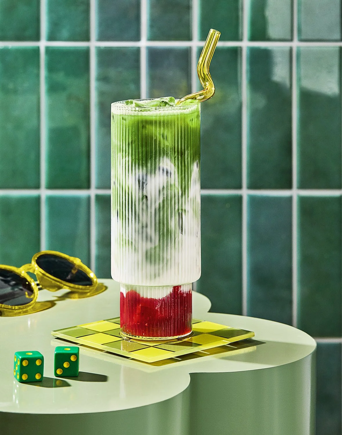  A tall ribbed glass filled with a layered iced strawberry matcha latte featuring bright green matcha on top, creamy white almond milk in the middle, and vibrant red strawberry fruit at the bottom, garnished with a bent yellow glass straw. The glass 