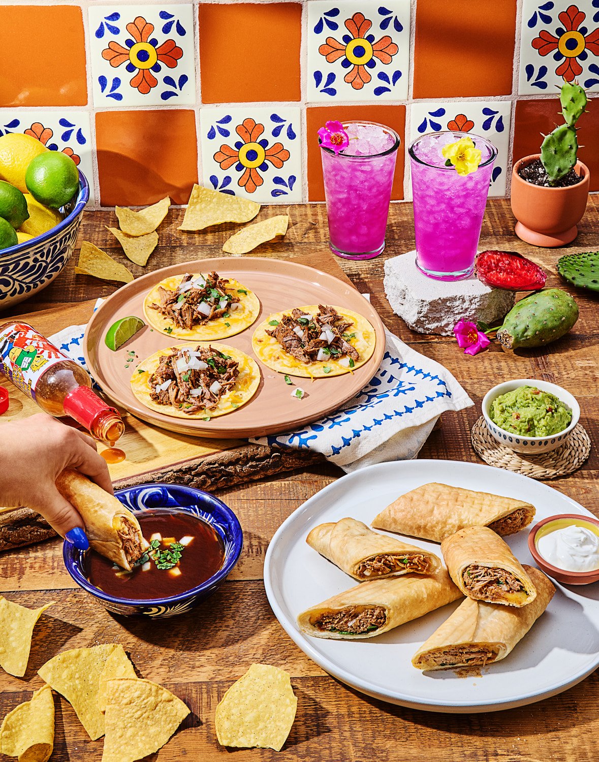  A colorful Mexican-inspired food spread on a rustic wooden table. In the center, three tacos topped with shredded meat, onions, and cilantro sit on a clay plate. Nearby, a hand dips a crispy birria flautas into a rich, dark consommé in a blue patter
