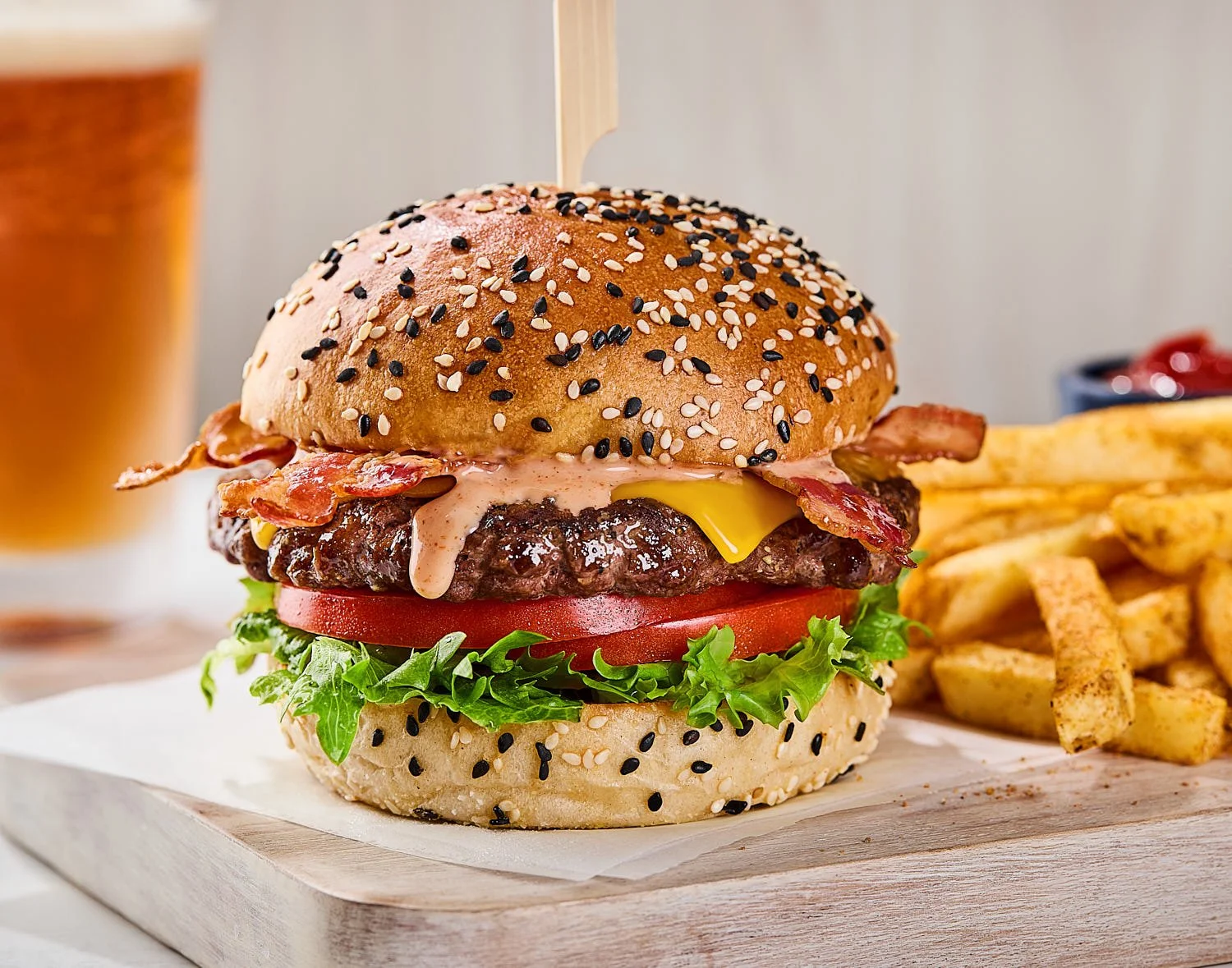 Juicy  bacon cheeseburger with lettuce and tomato slices on a cutting board. There is a sauce drip on the burger with fries and a beer in the background. Light and bright restaurant photography by Seattle based photographer Mike Gluckman.
