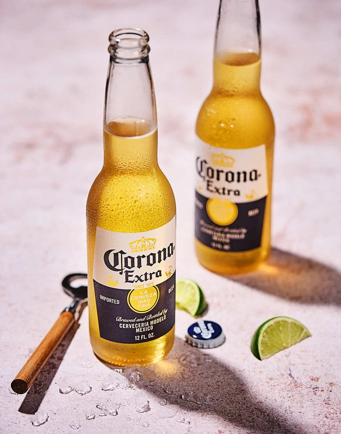 Menu image of two bottles of Corona beer, a bottle cap, lime wedges, and a bottle opener on a warm stone surface. Bright sunny lighting with crisp shadows. Created by Seattle restaurant photographer Mike Gluckman