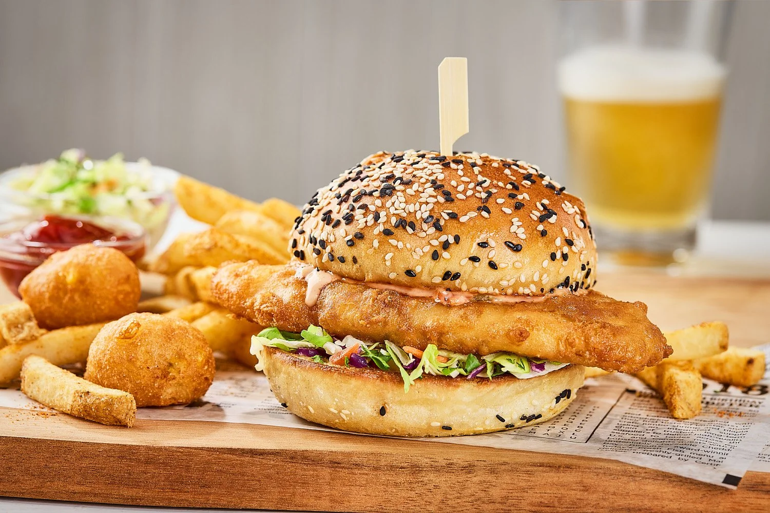 Crispy fried cod sandwich with coleslaw on a sesame seed bun on a board with fries and hush puppies. In the background is a draft beer. Light and bright with soft shadows. Seattle seafood restaurant photographer Mike Gluckman.