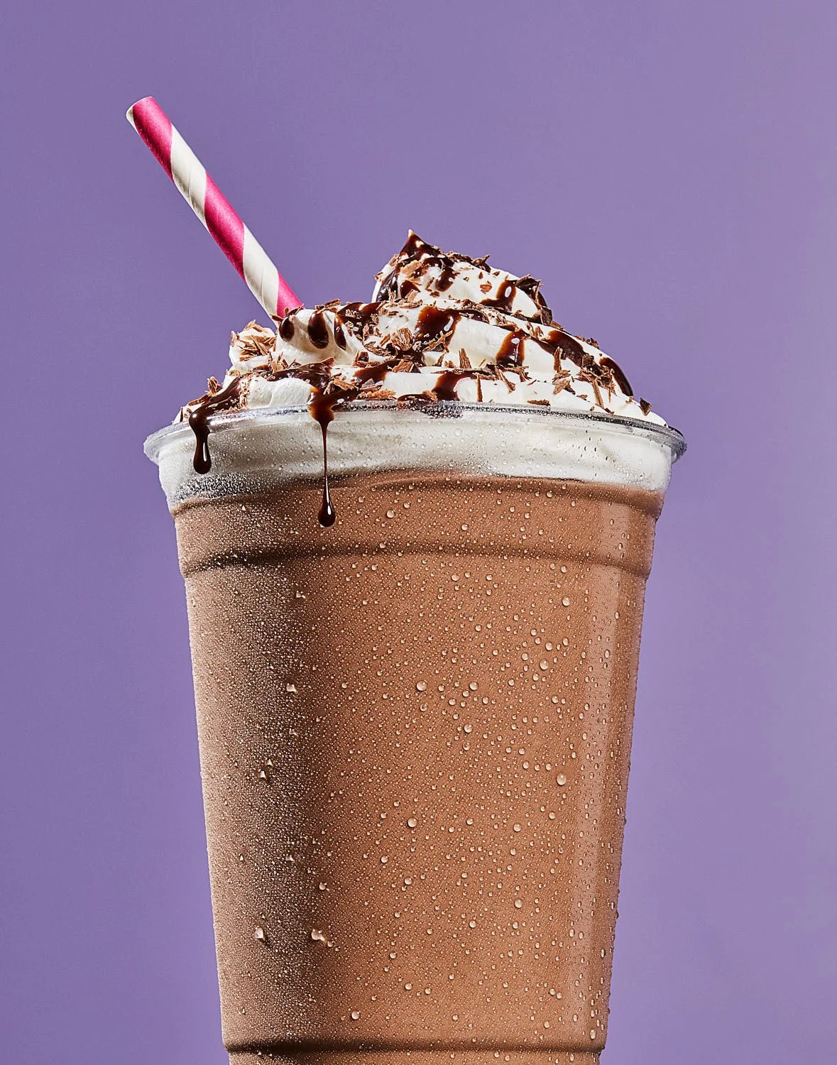 Close-up of a cold chocolate milkshake topped with whipped cream, chocolate shavings, and chocolate syrup, with a pink and white striped straw against a purple background. Milk shake photography by Seattle photographer Mike Gluckman.