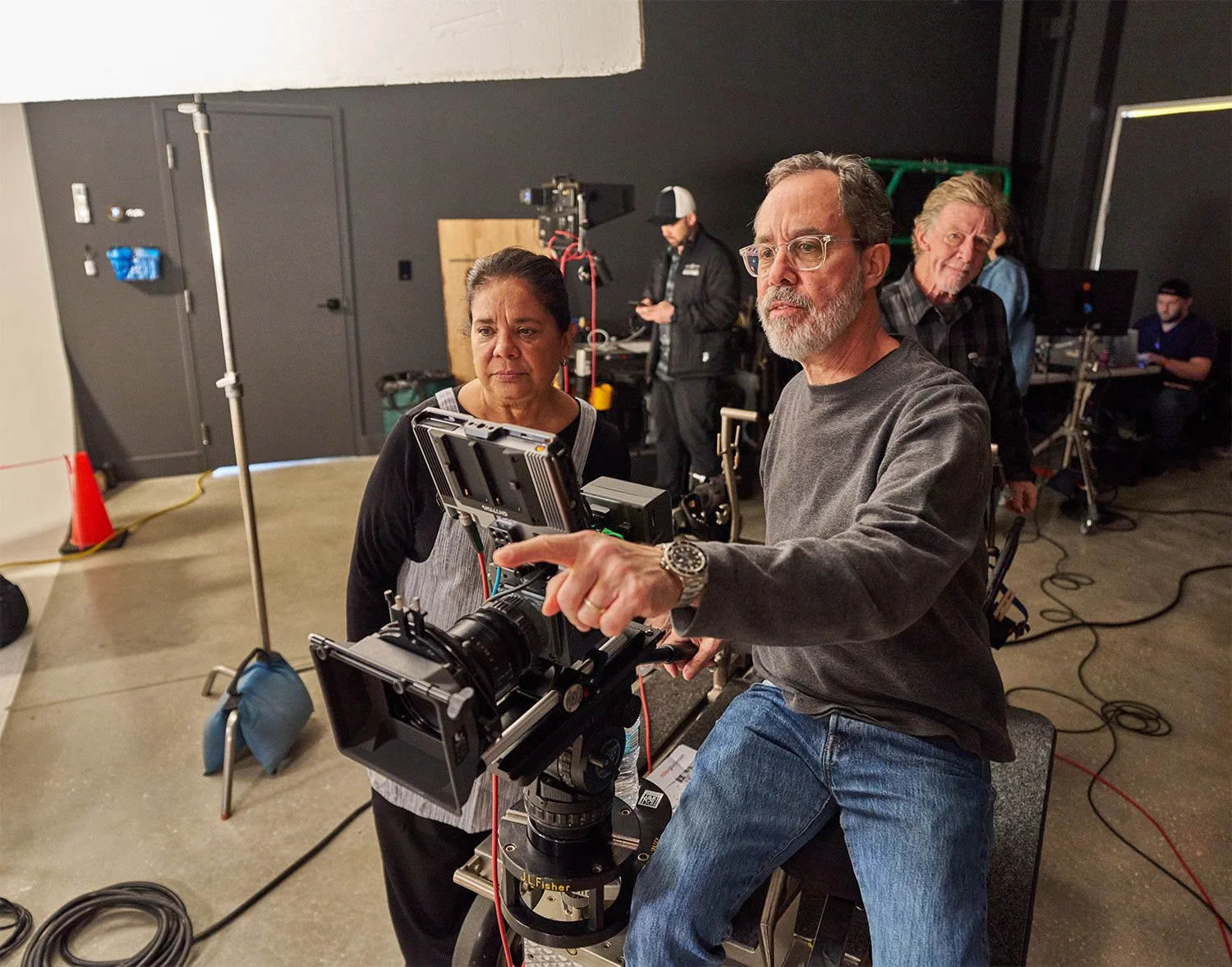 BTS of Seattle based food director & dp Mike Gluckman operating the camera while pointing to something on set for food stylist Aracelis Thompson. In the background are other crew members in the studio including the dolly grip and 1st assistant camera