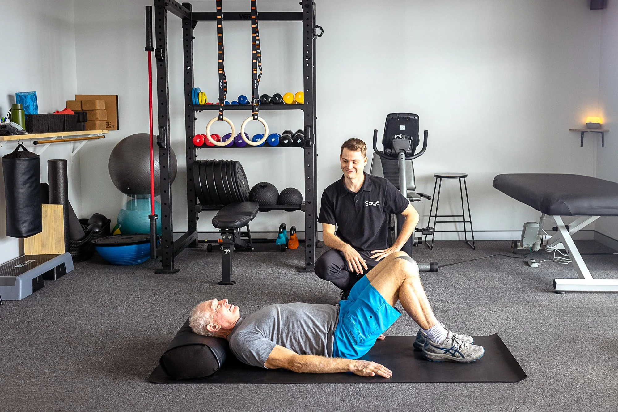 Older man exercising on a mat in a gym with Exercise Physiologist coaching him; exercise equipment in background.