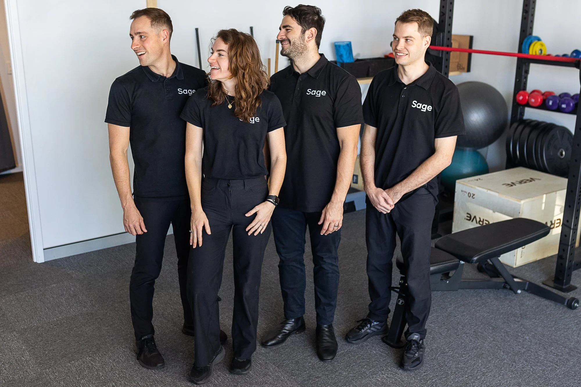 Four people wearing matching black polo shirts with "Sage" logo, standing in a gym setting with exercise equipment.