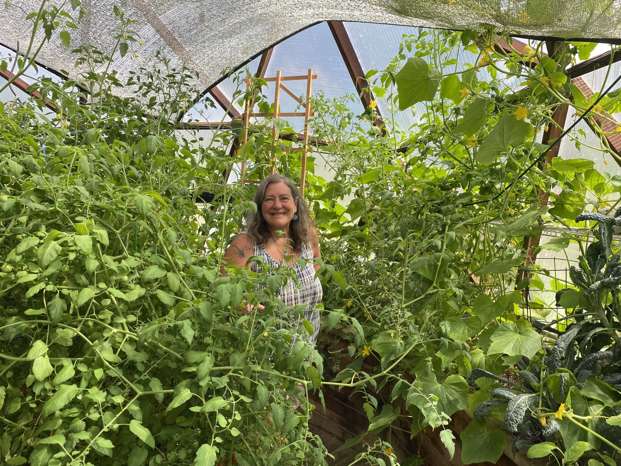 Woman with long hair smiling in a greenhouse filled with green plants and yellow flowers.