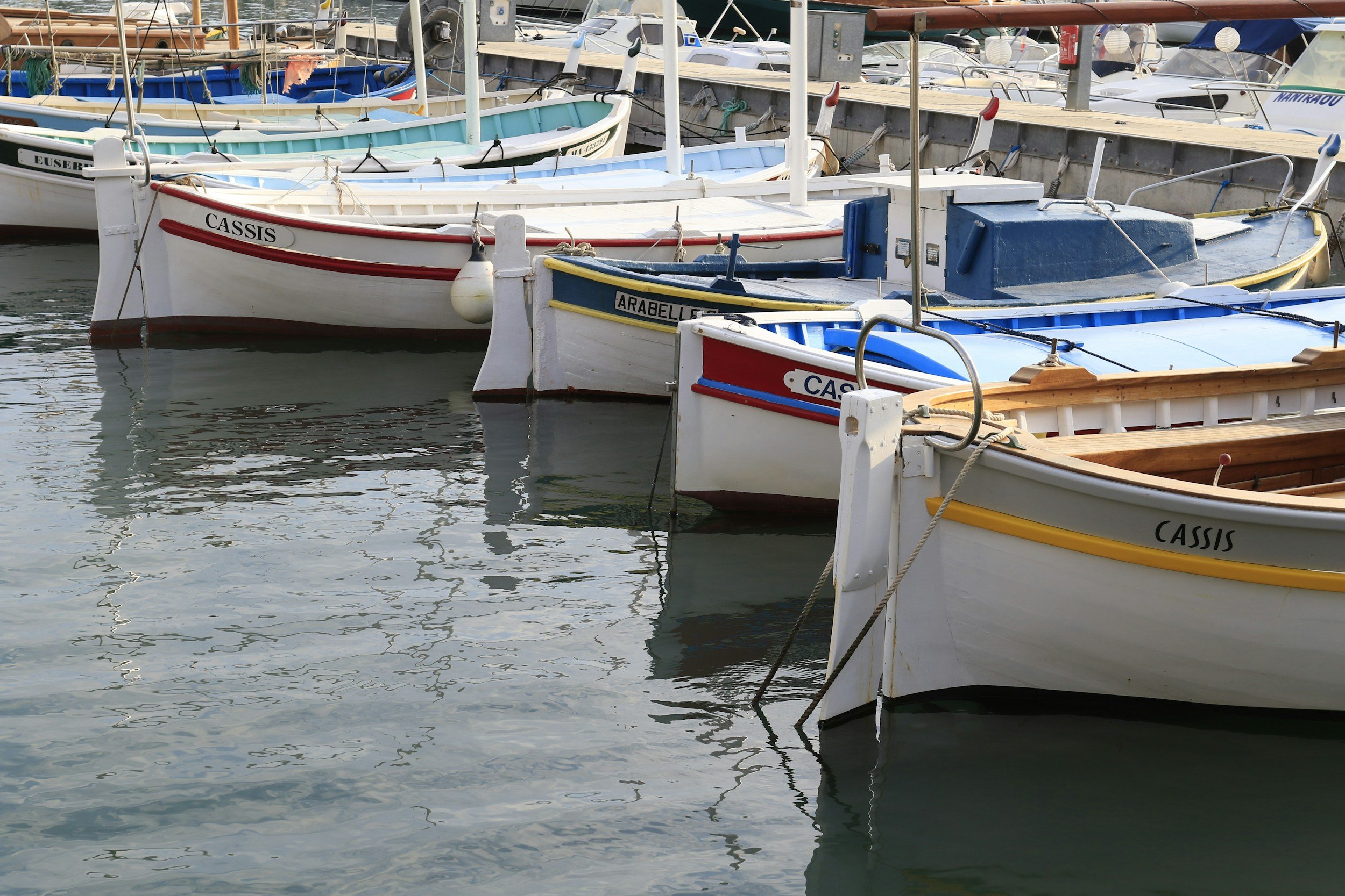 Multiple small boats docked at a marina, with their bow tags reading 'CASSIS' and other names, floating in calm water.