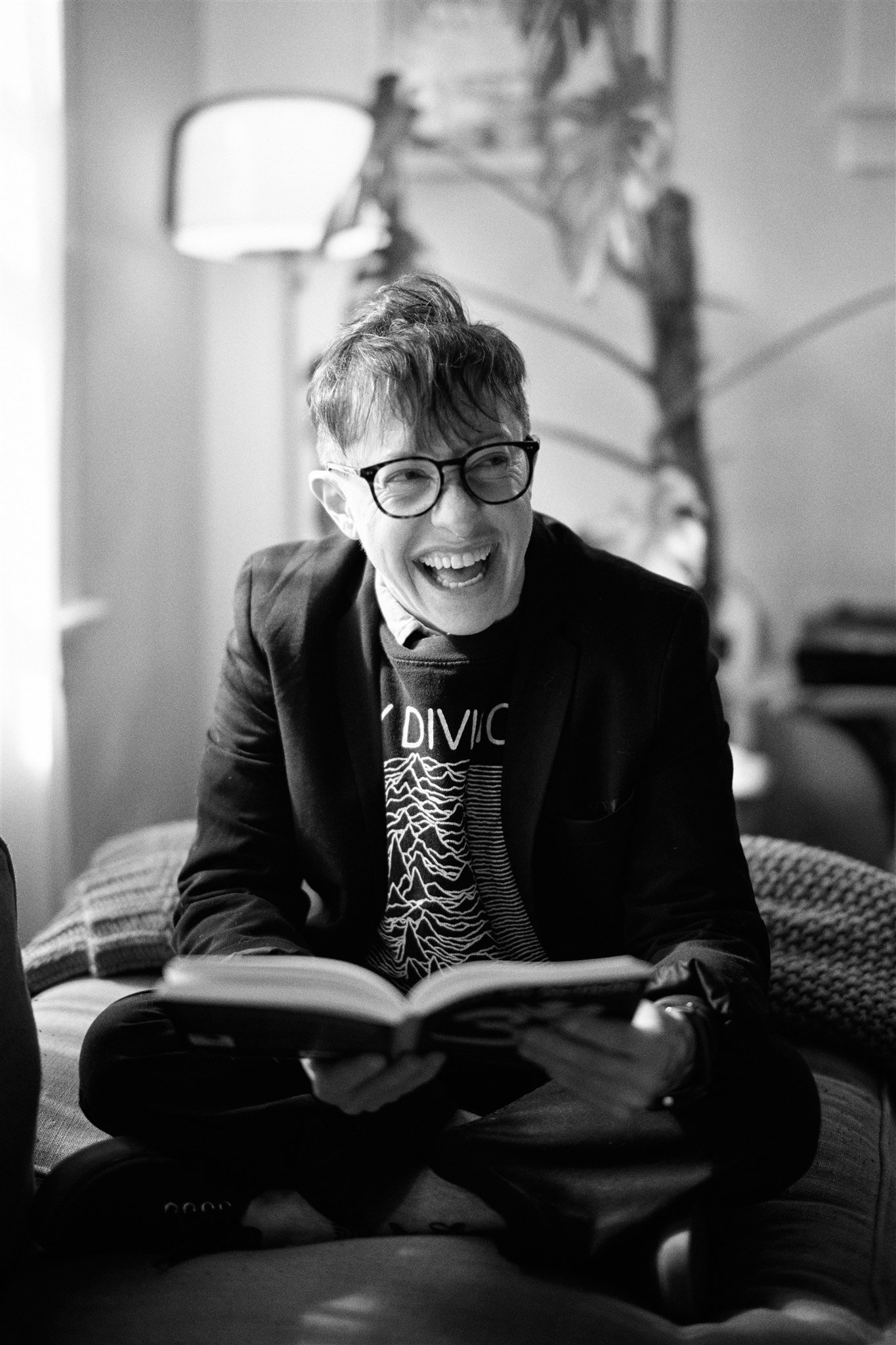 A black and white photo of a person with glasses laughing while sitting cross-legged on a sofa, reading a book, with a lamp and a bookshelf in the background.