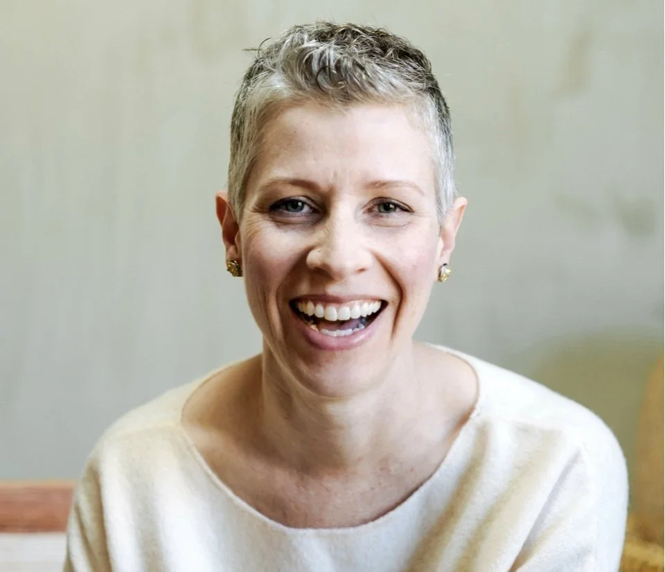 A woman with short, gray hair smiling, wearing earrings and a light-colored top, in front of a plain background.