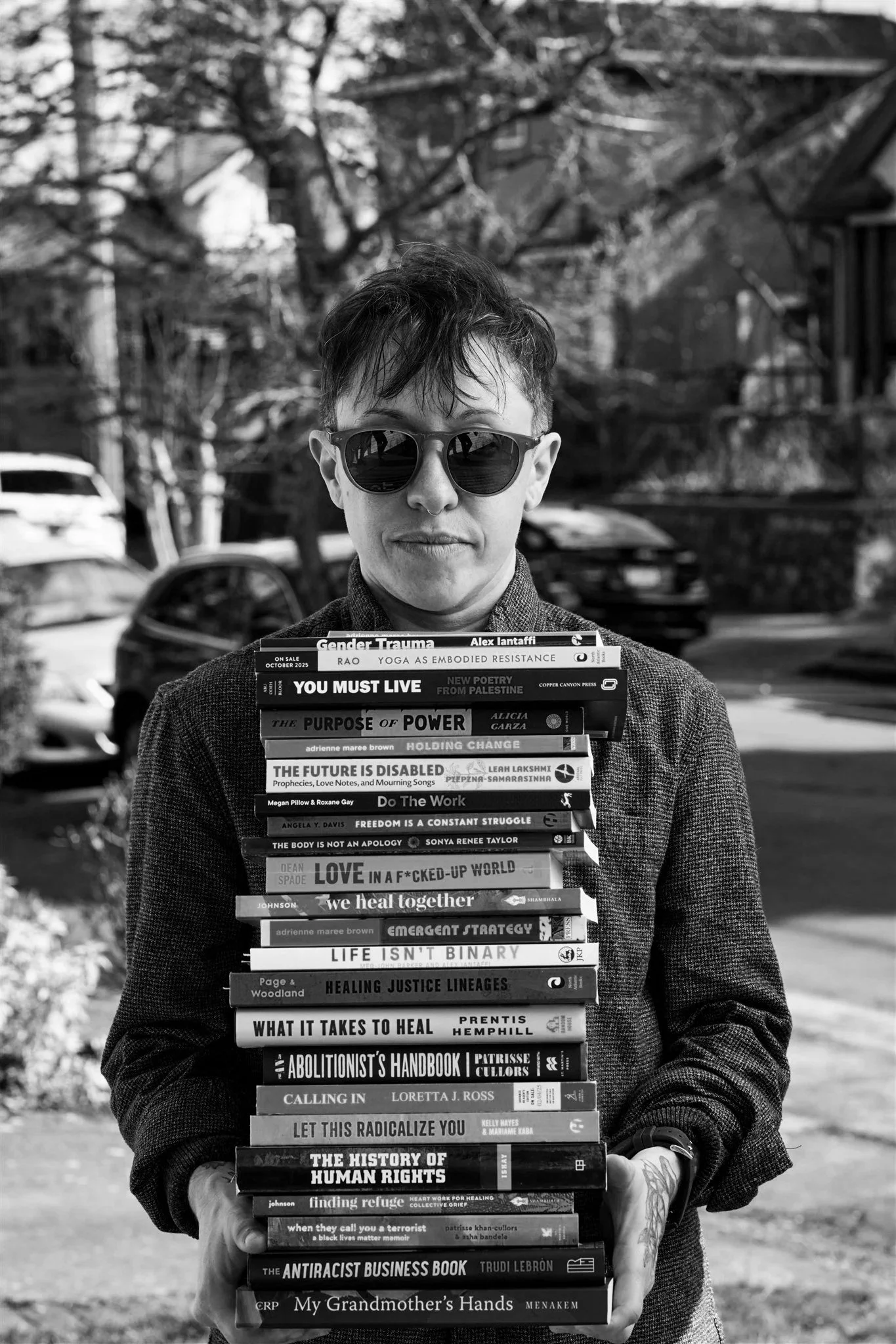 Black and white photo of a person wearing sunglasses, holding a tall stack of books with various titles, outdoors with trees and parked cars in the background.