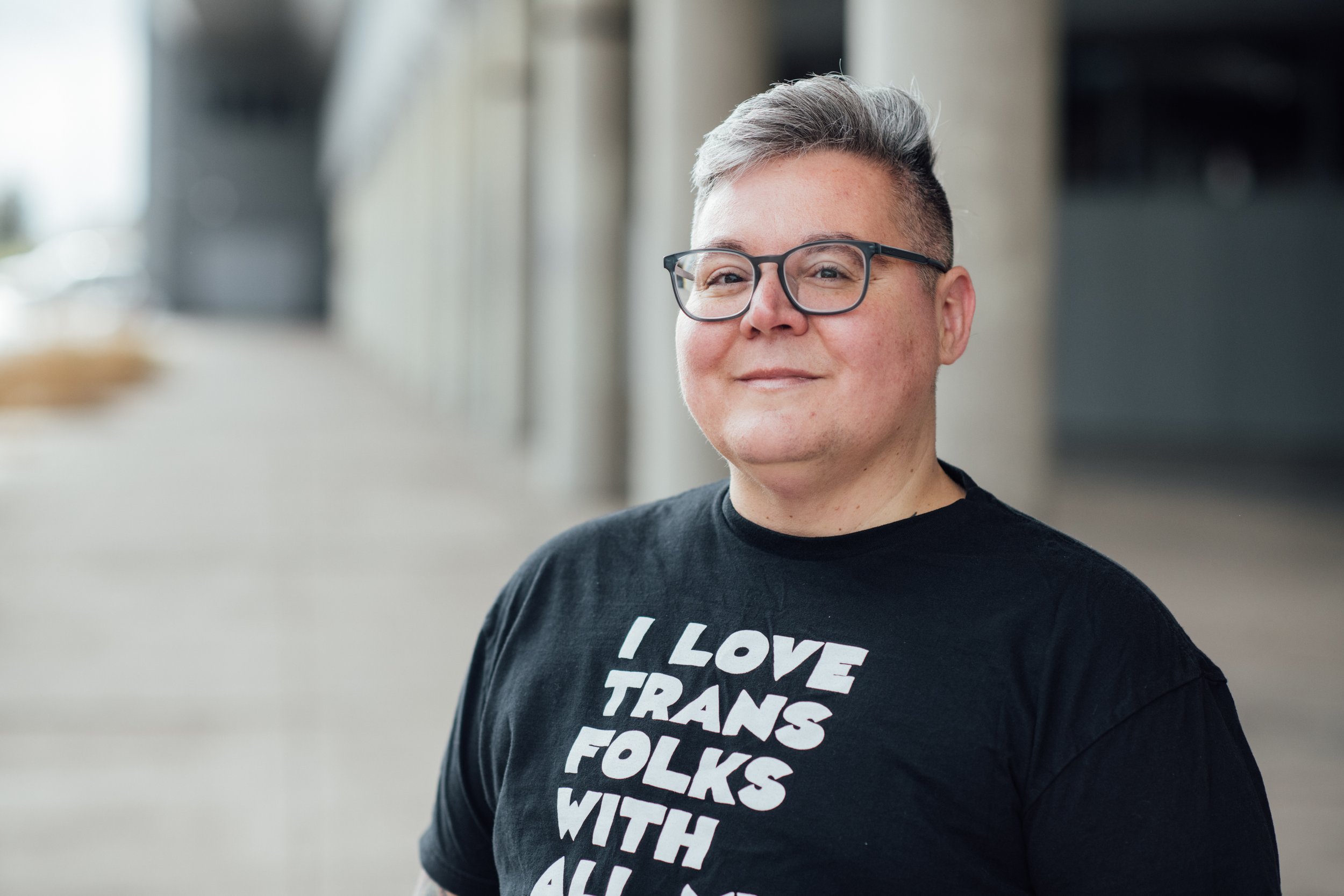A person with short gray hair, glasses, and a black T-shirt with white text. The background appears to be an indoor or outdoor public space.
