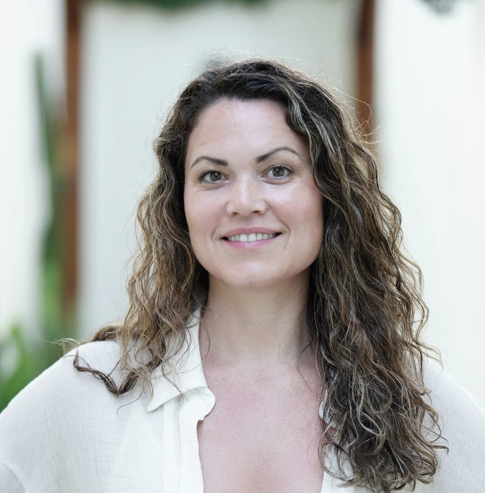 A woman with long curly brown hair wearing a white blouse, smiling gently, standing indoors with a blurred background.