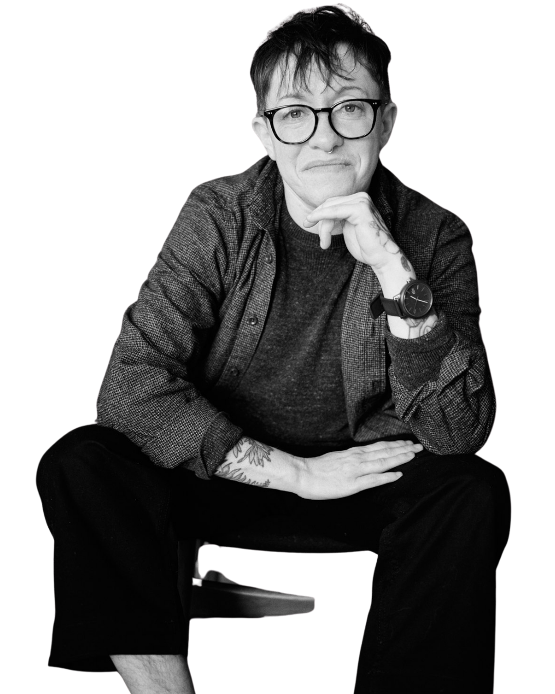 Black and white photo of a man wearing large glasses, a dark shirt, and a watch, sitting on a chair with one hand resting on his knee and the other hand supporting his chin.