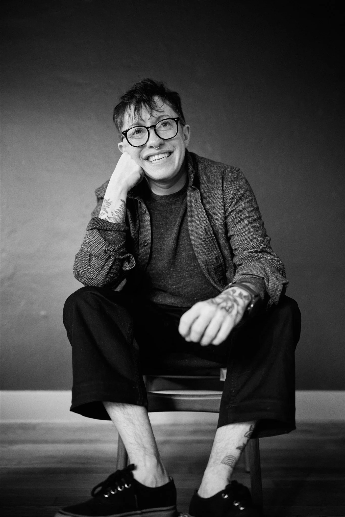 Black and white photo of a smiling person with short hair, glasses, and tattoos, sitting on a chair with one leg up, wearing a dark sweater, shorts, and sneakers, against a plain dark background.