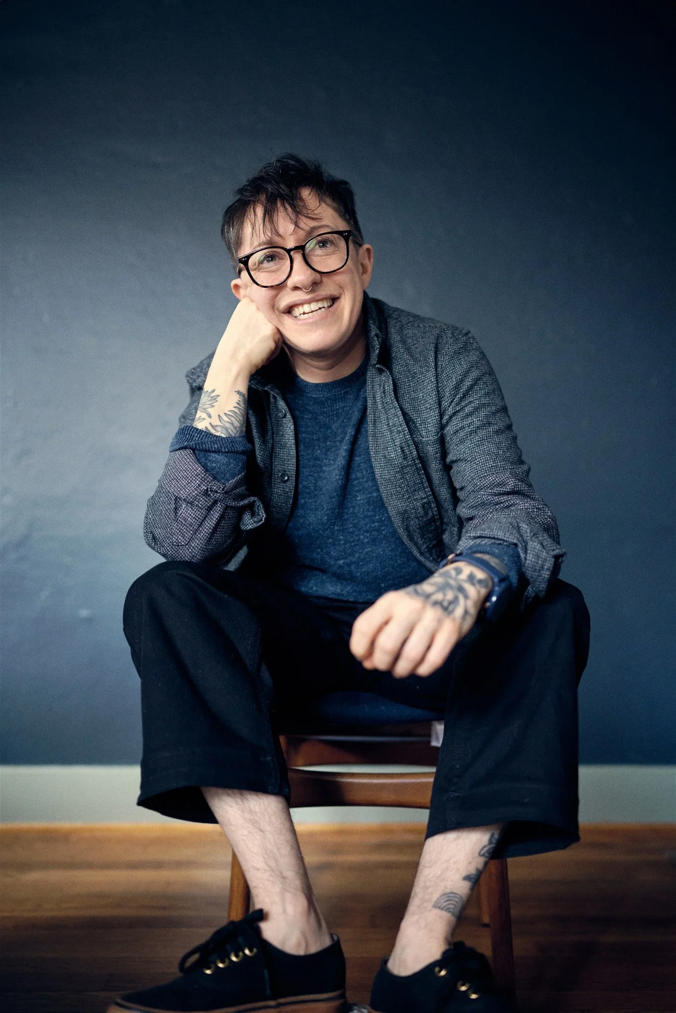 A young man with glasses, tattoos, and a septum piercing sitting on a wooden chair, smiling and resting his head on his hand, against a blue wall background.