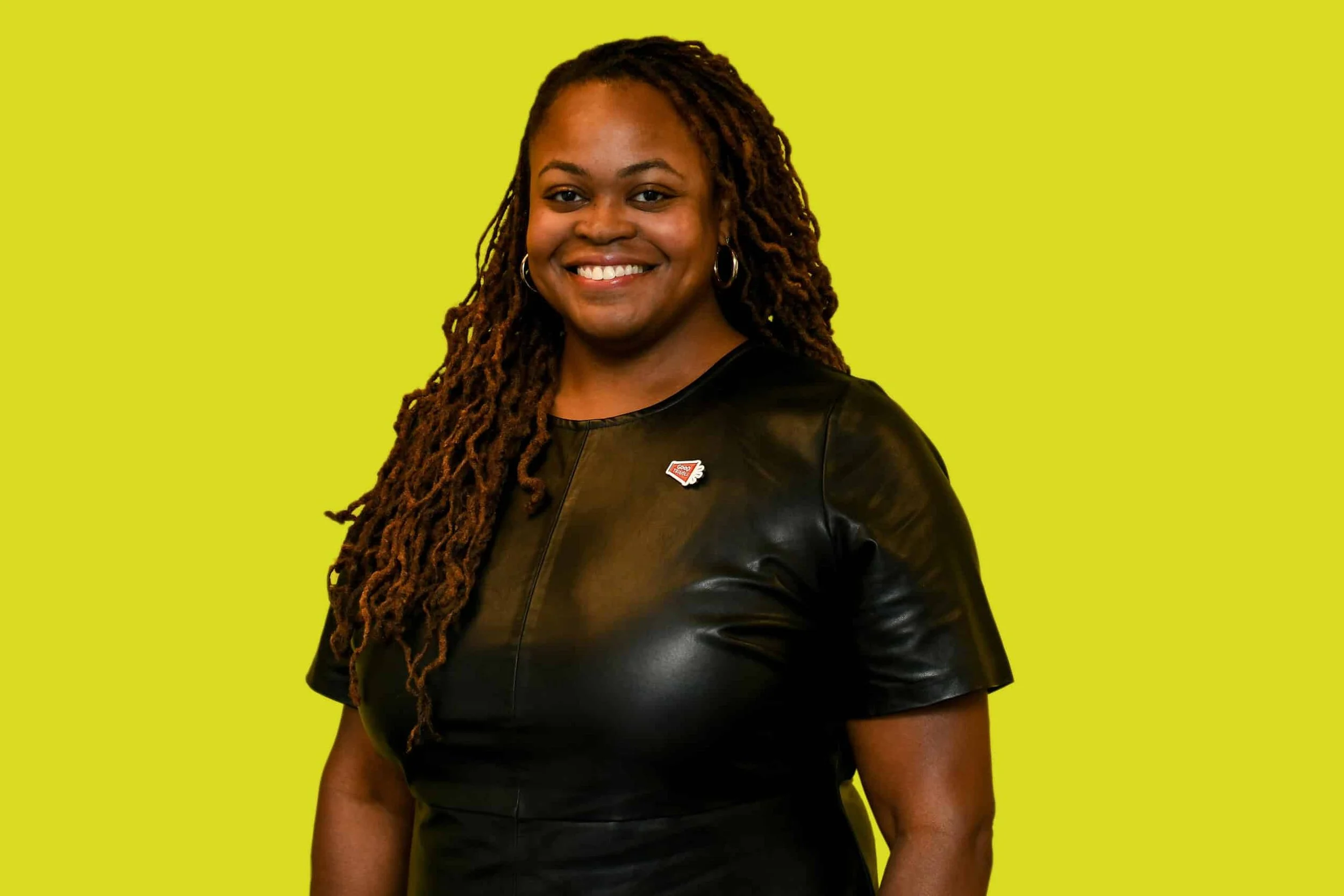A woman with long, styled dreadlocks smiling, wearing hoop earrings and a black leather top, standing against a lime green background.