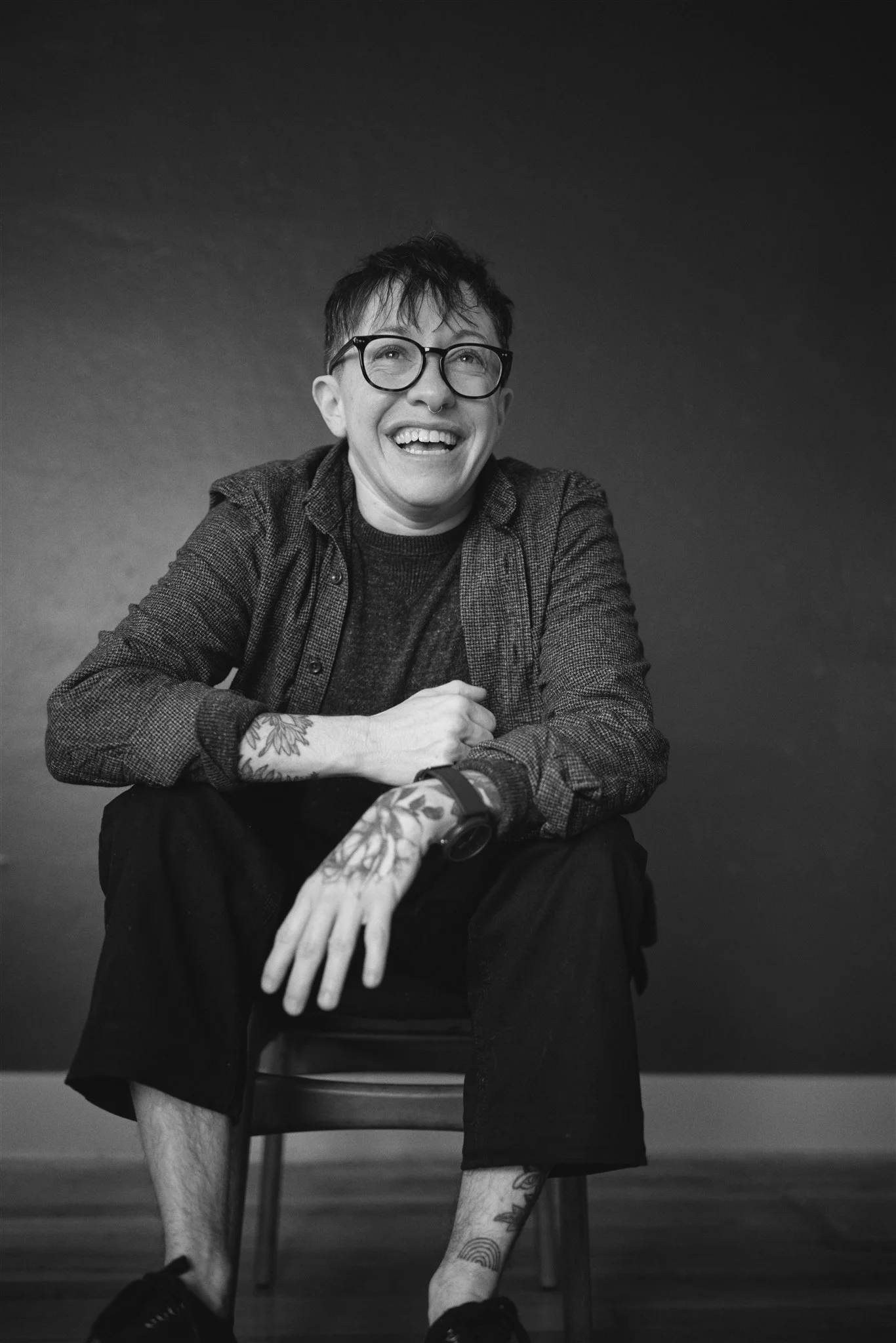 Black and white photo of a person sitting on a chair, smiling and looking upwards. They have short hair, glasses, a septum piercing, tattoos on their arms and legs, and are wearing a casual jacket and shorts.