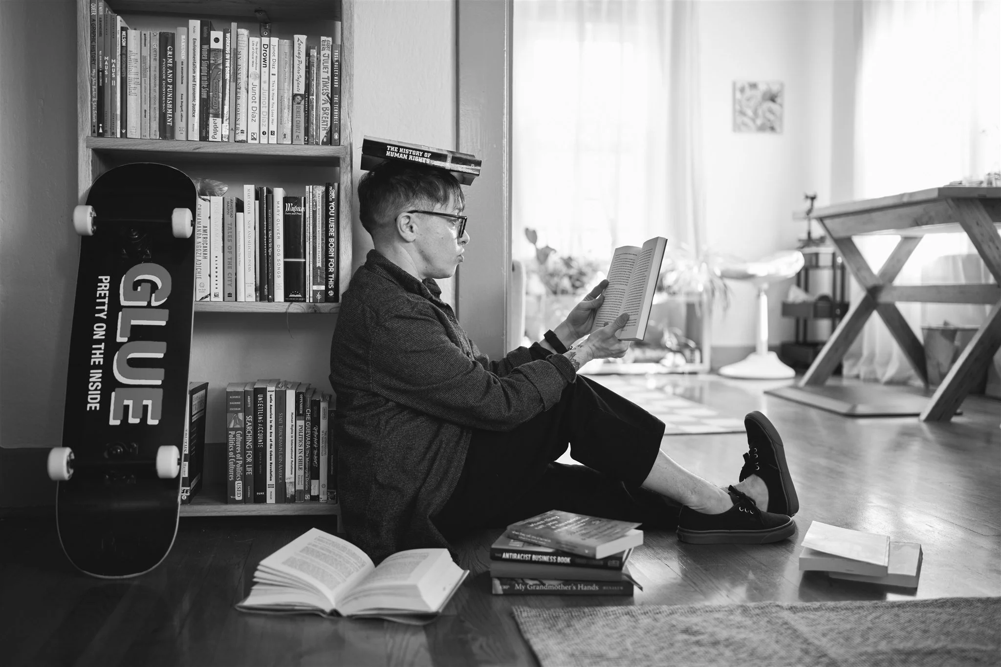 A person with short hair and glasses is sitting on the floor, reading a book. They are surrounded by several books on the floor and sitting in front of a bookshelf filled with books. A skateboard leans against the bookshelf. There are more books and a skateboard in the background and on a table near the window, which has curtains and natural light.