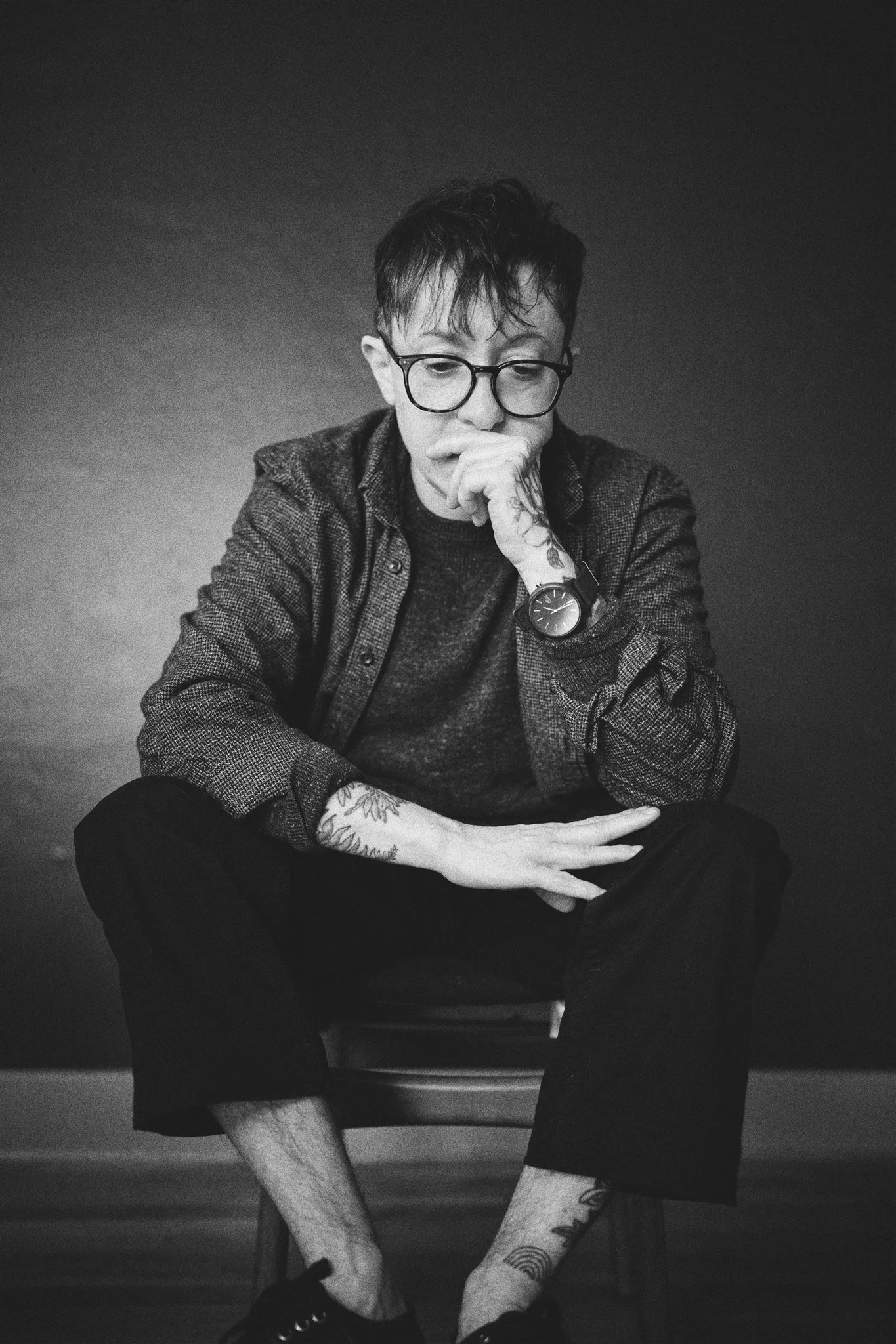 Black and white photo of a young man sitting on a chair with his legs crossed, resting his chin on his hand, wearing glasses, a watch, and casual clothing, with visible tattoos on his arm and legs.