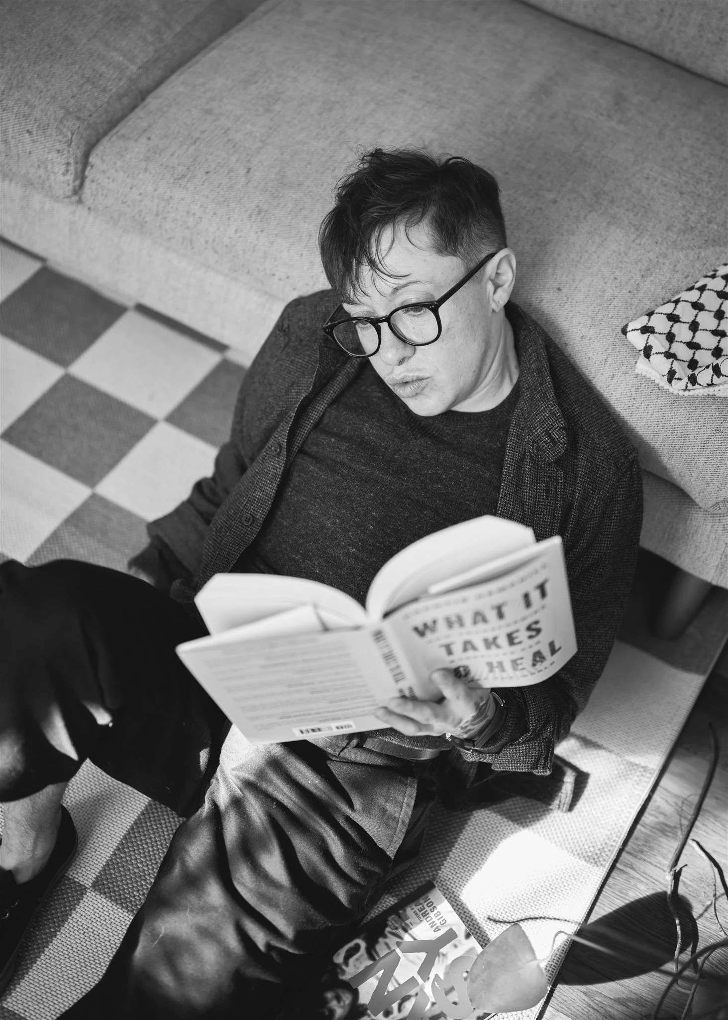 A person with glasses and dark hair is sitting on the floor, leaning against a sofa, reading a book titled 'What It Takes,' with a patterned pillow nearby and a bag on the floor. The photo is in black and white.